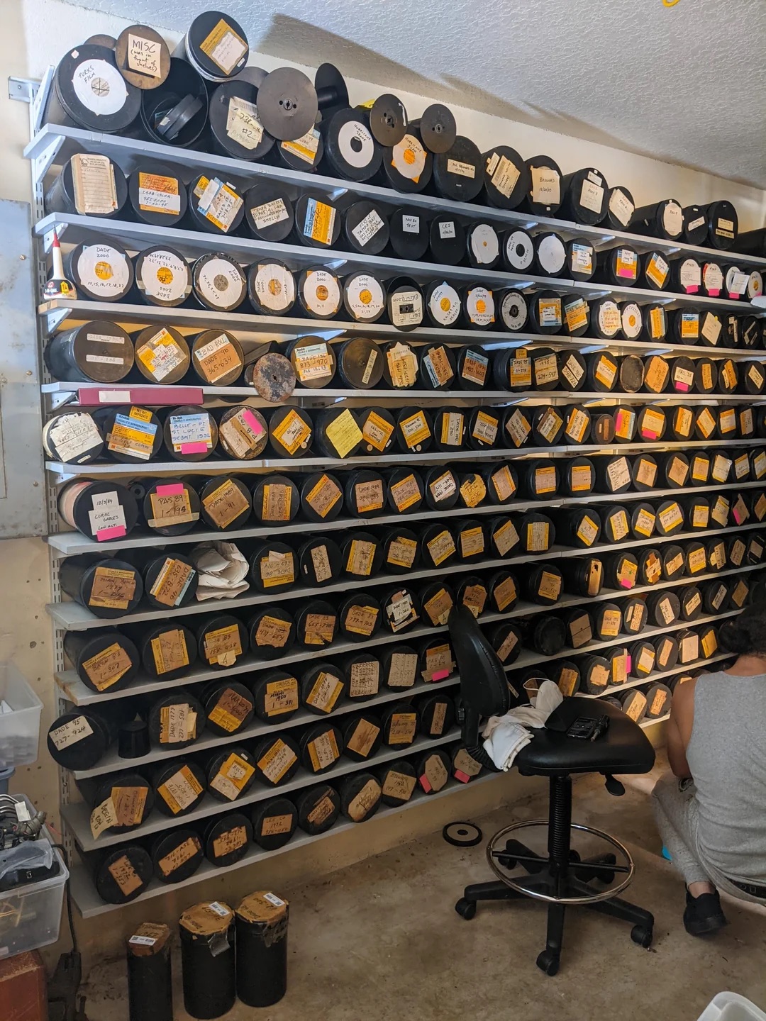 A large shelving unit filled with dozens of reels of film or tape, each individually labelled with handwritten or printed tags. The reels are neatly arranged, covering the entire wall from floor to ceiling. A person is seen sitting on the floor in the lower right corner, working with some of the materials. The scene suggests a substantial analogue media archive or restoration project.